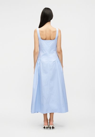 Γυναίκα που στέκεται με την πλάτη εμφανή, φοράει ένα ανοιχτό μπλε μίντι φόρεμα χωρίς μανίκια και λευκές πέδιλες με τακούνι, δίπλα σε απλό φόντο.