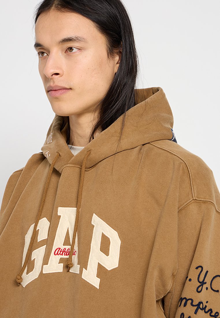 Braun-gefütterter Sweatshirt mit einem großen cremefarbenen "GAP"-Logo, roten Akzenten und navyblauem Text auf dem Ärmel. Baumwollmaterial mit weicher Textur.