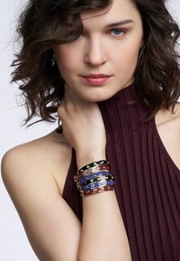 Bracelets colorés en couches arborant des bandes noires, rouges et bleues avec des accents en métal doré, portés au poignet contre un haut maroon texturé.