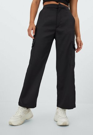 Woman wearing high-waist black wide-leg cargo pants with side pockets and chunky white sneakers, standing with hands relaxed by sides.