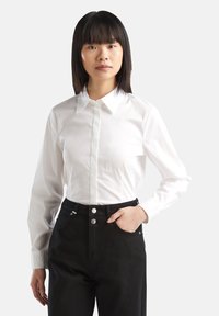 Camicia bianca con colletto a bottone e maniche lunghe, realizzata in tessuto morbido, abbinata a pantaloni neri a vita alta con bottoni argentati.