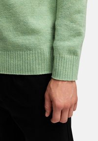 Myntgrön tröja i mjukt stickat material med ribbade ärmar. Fotot visar ärmen och en hand som vilar mot en mörk yta.