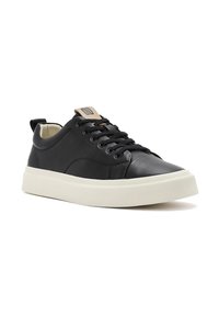 Sneaker in pelle nera con suola in gomma bianca, punta rotonda, design con lacci e un'etichetta in tinta ocra sulla linguetta. Include una piccola linguetta sul tallone.