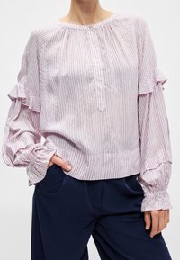 Femme portant un chemisier rose clair rayé avec des manches à volants et des boutons sur le devant, associé à un pantalon bleu marine foncé.