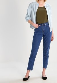 Veste bomber bleu clair sur un haut vert foncé, associée à un jean ajusté taille haute bleu foncé et des chaussures plates noires. Tissu lisse et coupe ajustée.