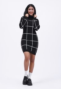 Melna turtlenecka kleita ar baltu režģa rakstu, pieguļošs dizains, izgatavota no mīksta materiāla. Modelis valkā melnas kurpes ar baltām zeķēm.