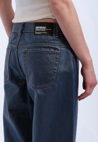 Mörka denimbyxor har hög midja, raka ben och kontrasterande gula sömmar. Bakfickorna är synliga, med en logotagsetikett.