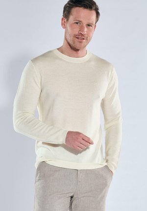 Man wearing a cream long-sleeve sweater and light gray pants, standing against a plain light background.