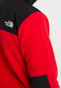 Giacca in pile rossa con pannello in nylon nero sulla spalla. Il logo The North Face è stampato in argento sulla parte superiore sinistra.