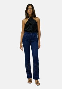 Femme portant un haut noir à bretelles, un jean taille haute bleu foncé avec des motifs brodés près des chevilles, et des sandales noires à lanières.