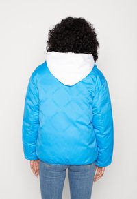 Blue puffer jacket with white hood, featuring subtle all-over branding "TOMMY JEANS." Smooth, shiny fabric, relaxed fit, and ribbed cuffs.