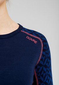 Marineblå langærmet top med et geometrisk blåt mønster på skulderen, accentueret med røde stikninger og en struktureret overflade. Mærket læser "DOVRE."
