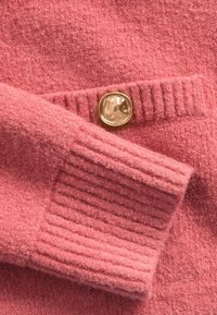 Rózsaszín kötött pulóver, texturált felülettel, bordázott mandzsettákkal és az elején fényes, kerek arany gomb díszítéssel.