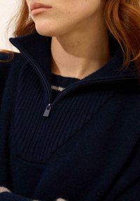 Granatowy sweter o prążkowanej fakturze, z kołnierzem na pół zamek i kontrastową, paskowaną wewnętrzną warstwą. Materiał wydaje się miękki i ciepły.