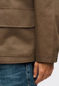 Καφέ μανίκι μπουφάν και τσέπη με λεπτομέρεια από κουμπί πάτημα, φορεμένα με μπλε τζιν τζιν, χέρι ακουμπισμένο στο πλάι.