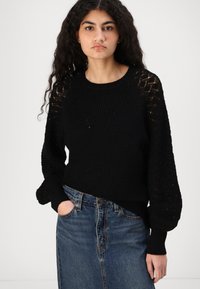 Maglione nero lavorato a maglia con maniche testurizzate e motivi geometrici; abbinato a una gonna di jeans blu, che mette in evidenza una vestibilità rilassata.
