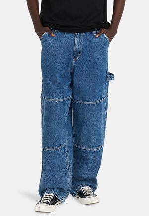 Blauwe denim cargobroek met een losse pasvorm, grote zijzakken en ingewikkelde stikdetails. Gecombineerd met zwarte sneakers en een zwart shirt.