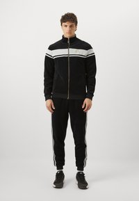 Svart velour treningsdrakt med full glidelåsjakke med hvite og gull detaljer, matchende bukser med hvite sidestriper og elastiske mansjetter.