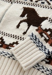 Knitted zip sweater in cream, featuring brown horse motifs and geometric patterns. Ribbed cuffs with navy detailing. Soft texture.