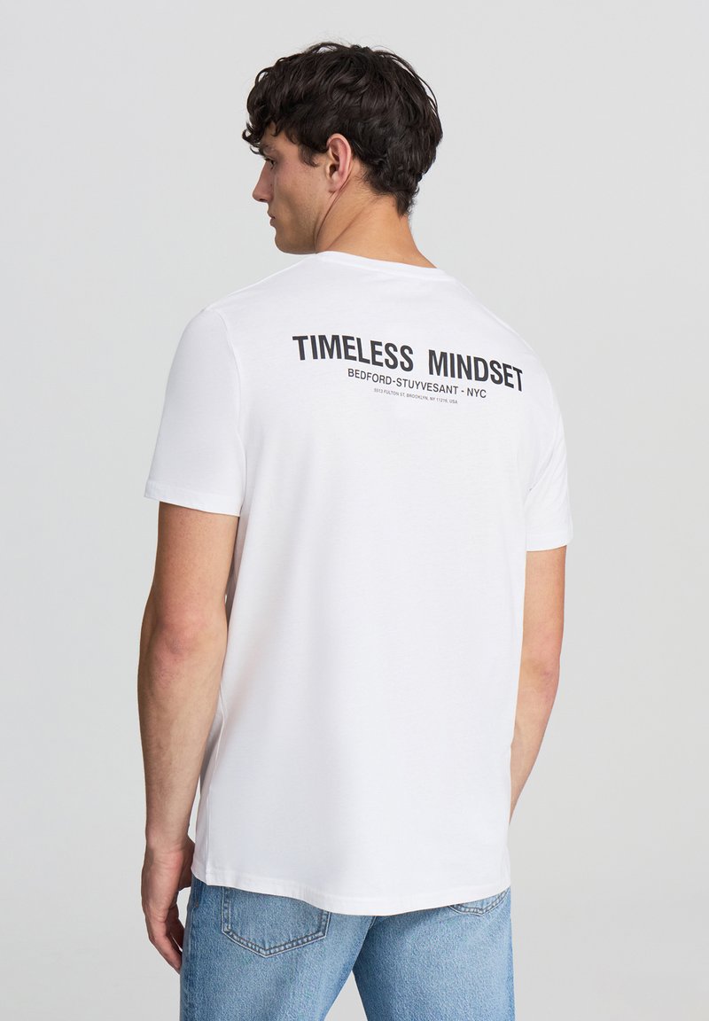 Biała bawełniana koszulka z okrągłym dekoltem, krótkim rękawem oraz czarnym napisem na plecach "TIMELESS MINDSET" i szczegółami lokalizacji.