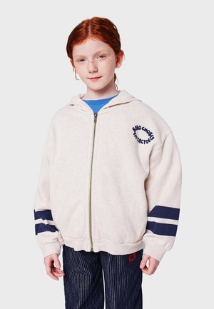 Cremfarbener Reißverschluss-Hoodie aus weichem Stoff, mit einem navyblauen Druck auf der linken Brust und gestreiften Akzenten an den Ärmeln. Getragen über einem blauen Oberteil.
