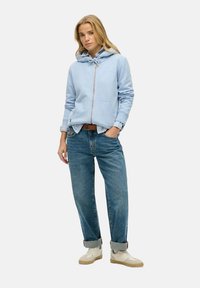 Felpa azzurro chiaro con zip e cappuccio regolabile, indossata sopra una maglietta bianca, abbinata a jeans blu con risvolti e sneakers beige.