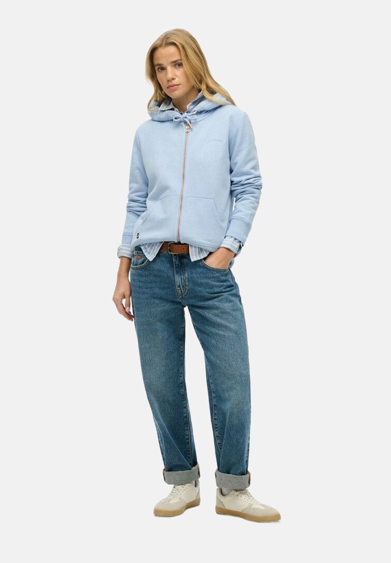 Felpa azzurro chiaro con zip e cappuccio regolabile, indossata sopra una maglietta bianca, abbinata a jeans blu con risvolti e sneakers beige.