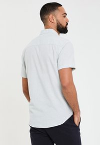 Camisa corta de manga corta color gris claro con cuello abotonado, textura suave, dobladillo curvado y detalle en el yugo trasero, combinada con pantalones oscuros.