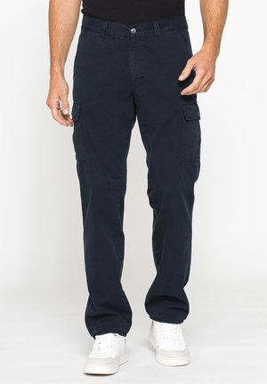 Uomo che indossa pantaloni cargo blu scuro con tasche con patta e sneaker bianche, in piedi su uno sfondo bianco semplice.