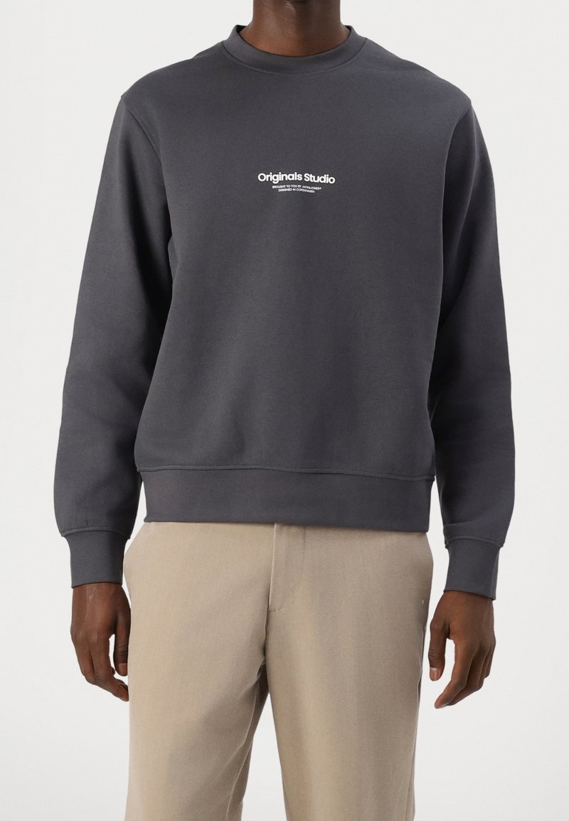 Homme portant un sweat-shirt gris foncé à col rond avec un petit texte blanc "Originals Studio" et un pantalon beige, debout devant un fond clair.
