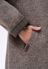 Houndstooth wollen jas met een groene geruite manchet, voorzien van een zijzak en een bruine label met de tekst "blue shadow." Textuur van het breipatroon zichtbaar.