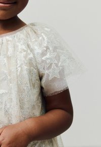 Petite fille portant une robe argentée métallique transparente avec des motifs d'étoiles, des manches bouffantes et une superposition en tulle texturé. Sa carnation est foncée.