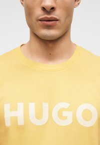 Νεαρός που φοράει κίτρινο μπλουζάκι με λευκό κείμενο "HUGO" στο στήθος, εμφανιζόμενος από τον λαιμό μέχρι το πάνω μέρος του κορμού.