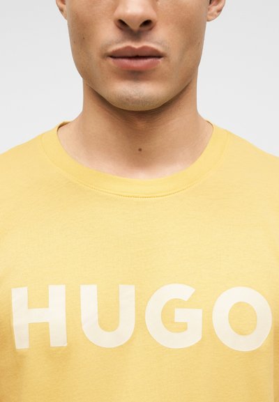 Млад човек, носещ жълта блуза с бяла надпис "HUGO" на гърдите, показан от врата до горната част на торса.
