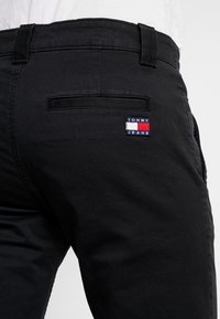 Černé džíny s zadní kapsou, na které je vyšitá značka "Tommy Jeans" v červené, bílé a modré barvě, a hladkým povrchem.