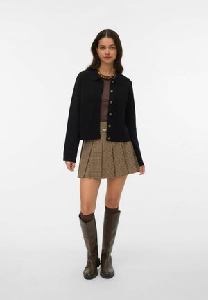 Veste noire courte à boutons sur le devant, associée à une jupe plissée marron à carreaux et des bottes en cuir marron montant jusqu'aux genoux.