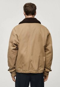 Beżowa kurtka z brązowym, teksturowanym kołnierzem z polaru, luźny krój, elastyczny pas oraz widoczne detale szwów na plecach.