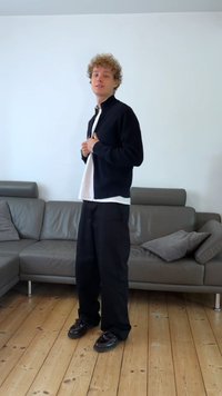 Jacket preto com fecho, sobre uma camisa branca, combinado com calças largas pretas e sapatos pretos, de pé em chão de madeira, contra uma parede branca.