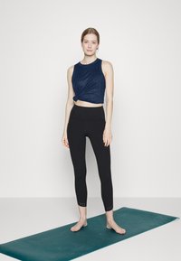 Top cropped azul-marinho com detalhe em nó, combinado com leggings pretas com pequenos recortes na barra, sobre um tapete de ioga teal.
