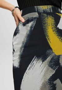 Main portant un bracelet en argent reposant dans la poche d'une jupe avec un motif abstrait de coups de pinceau noirs, blancs, gris et jaunes.