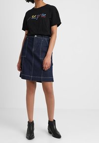 Camiseta negra con letras coloridas, combinada con una falda de mezclilla oscura. La falda tiene bolsillos delanteros y costuras visibles. Botas negras hasta el tobillo.