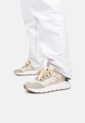 Baskets avec un mesh beige et des détails en suede ; accents verts et violets. Lacets blancs, semelle épaisse, embout rond. Portées avec un pantalon blanc ample.