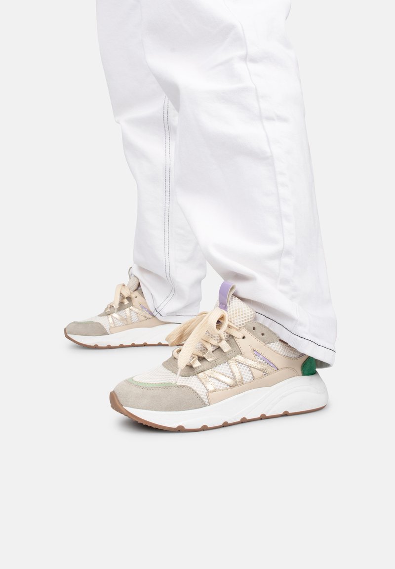 Baskets avec un mesh beige et des détails en suede ; accents verts et violets. Lacets blancs, semelle épaisse, embout rond. Portées avec un pantalon blanc ample.