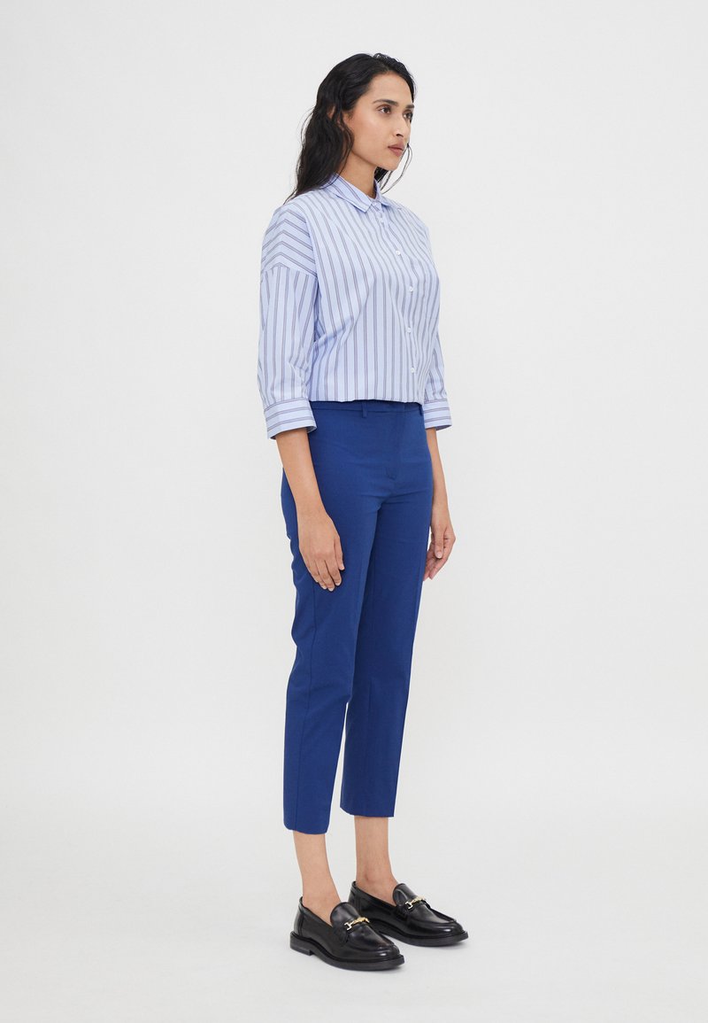 Chemise bleu et blanche à rayures avec des manches retroussées, assortie à un pantalon bleu ajusté et des mocassins noirs avec des accents dorés.