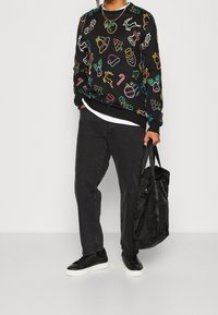 Schwarzer Pullover mit bunten, festlichen Designs, kombiniert mit schwarzen Hosen und schwarzen Sneakers. Enthält eine schwarze Tragetasche.