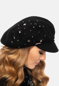 Cappello di lana nera decorato con elementi argentati, tra cui borchie e ciondoli, caratterizzato da una forma arrotondata e un ampio bordo.