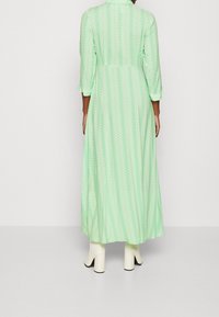 Robe longue vert clair à motif chevrons blancs, manches trois-quarts, taille ajustée, portée avec des bottes blanches à talons.