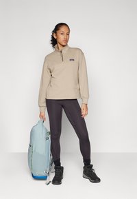 Beige fleece pullover met kwart rits en logo, gecombineerd met zwarte legging. Model houdt een lichtblauwe rugzak vast en draagt zwarte wandelschoenen.