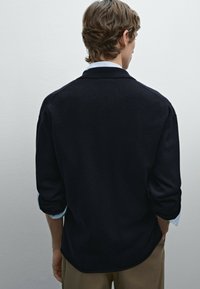 Navy knit sweater with a relaxed fit, featuring a collar and three-quarter sleeves. Smooth texture with rolled cuffs, layered over a light blue shirt.