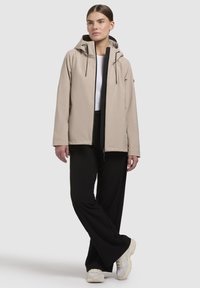 Beige jacka med huva, långa ärmar, dragkedja fram och svarta detaljer. Modellen bär svarta byxor och ljusa sneakers.